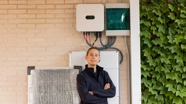 a person in a dark jacket crossing their arms and smiling in front of a solar battery system