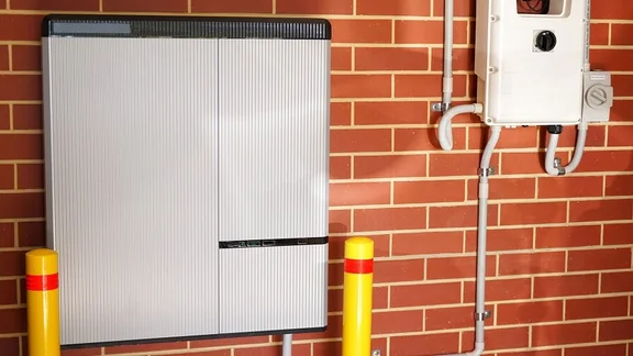Solar battery attached to a red-brick garage wall. There is an inverter to the right of the battery.