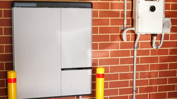Solar battery attached to a red-brick garage wall. There is an inverter to the right of the battery.