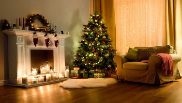 Warm and festive setting in a home with a decorated Christmas tree, three stockings above a fireplace, with assorted candles illuminating the fireplace