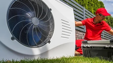 outdoor unit of a heat pump being worked on by a technician in red uniform