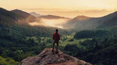 Man standing on cliff edge overlooking mountains and green valley
