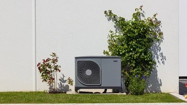 air source heat pump unit outside house with white wall, grass lawn, and shrubs