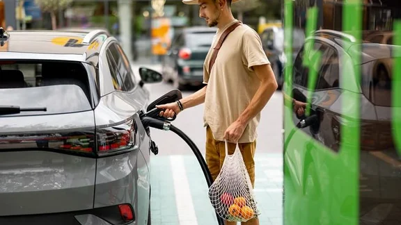 Man with groceries waiting for electric car to be charged on public charging station near a shopping area