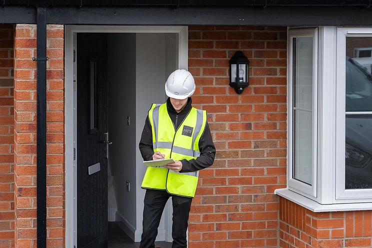 A heat battery installer stands outside a house with a clipboard.